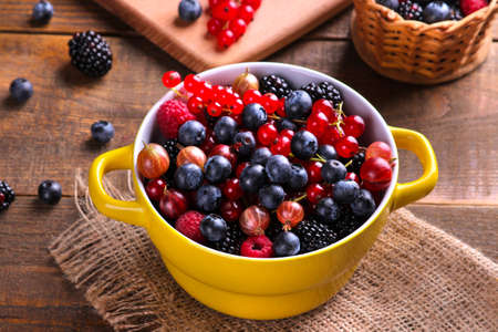 Various fresh berries including blueberries, raspberries, blackberries currants and gooseberries in a yellow bowl on a brown wooden background.の写真素材