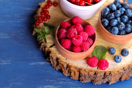 Fresh, large blueberries, currants and raspberries in bowls close-up on a wooden stand with mint stamens on a blue wooden background.の写真素材