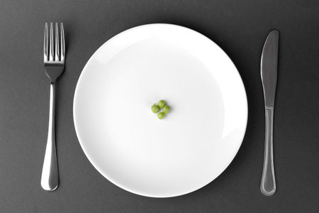 a few green fresh peas on a white plate next to a fork and knife on a black background. The concept...