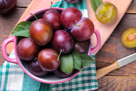 Fresh ripe plums with leaves in a bowl on a brown wooden table. top viewの写真素材