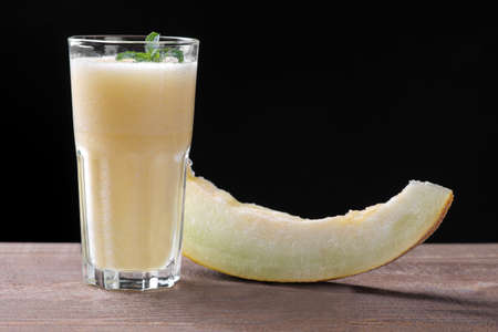 Smoothies of melon next to a slice of melon on a brown wooden table on a black backgroundの写真素材