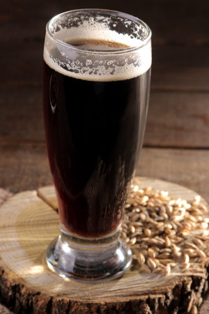 A glass of dark beer on a wooden stand with barley grains on a brown wooden table