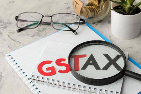 The word GST TAX under a magnifying glass with a notebook, glasses, pen and coins in a bank on a light concrete background.の写真素材