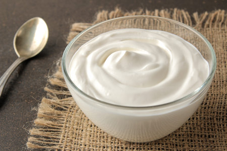 Greek yogurt in a glass bowl next to a spoon on a dark background. healthy food. natural yoghurt....