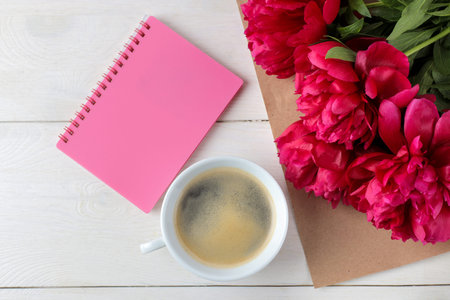 Beautiful bright pink flowers peonies and a notebook with a cup of coffee on a white wooden...