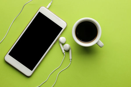 Smartphone White phone and headphones and a cup of coffee on a bright green background. top view.