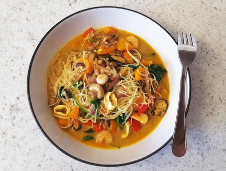 Rice noodles with fried red onion, baby mushrooms, red pepper, cooked with coconut milk, mussels, squid, king prawns, chopped garlic, fresh spinach, curry powder, and other spices.の写真素材