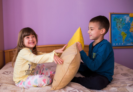 Brother and sister having fun in a pillow fight. Little boy is holding a pillow, while the girl  hits him with her pillow. Both are wearing their pajamas  Happy children in a pillow fightの写真素材