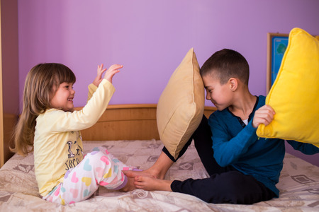 Brother and sister having fun in a pillow fight. Little boy is holding a pillow, while the girl  hits him with her pillow. Both are wearing their pajamas  Happy children in a pillow fightの写真素材