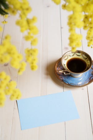 Cup of coffee and blue empty card on light wooden table and blooming mimosaの写真素材