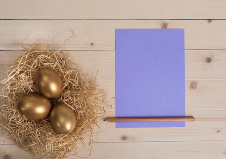 Three golden eggs in the nest and empty lilac card with pencilの写真素材