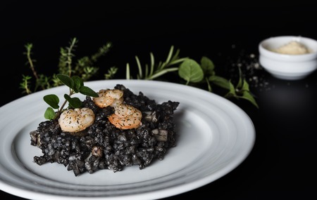 Plate with black risotto on black background with dramatic side light, aroma herbs and grated parmesan around plateの写真素材