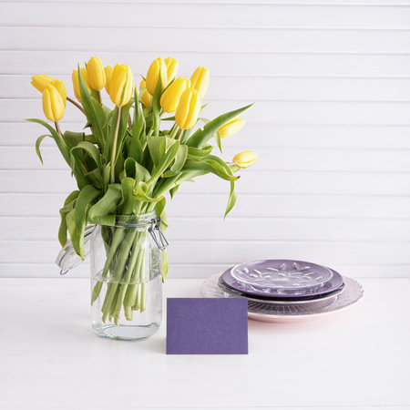 Yellow tulips, plates and empty card on white wooden table