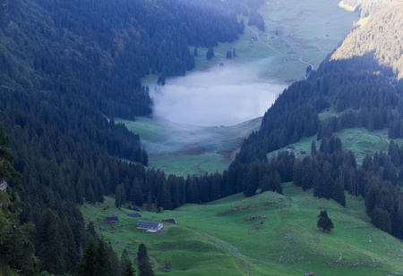 deep green valley with lake and mist floating on it and houses surounded by high,steep, rocky mountain walls at Säntis, Switzerlandの写真素材