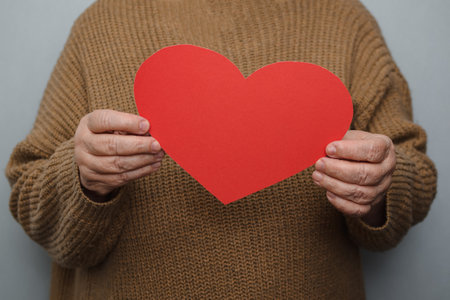 An elderly woman in a brown knitted pullover holds with both hands pointing at the camera a homemade red heart cut out of cardboard, expressing her love, support and care.の写真素材