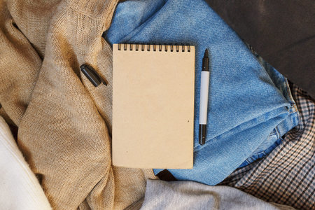 Flat lay top view on the table is a pile of clothes made of jeans, a knitted sweater, plaid trousers and a sweatshirt, an empty notepad with a marker lies on top, copy space.の写真素材