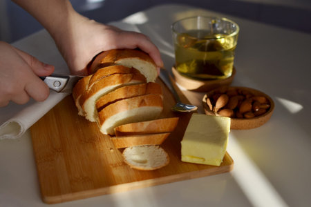 Early in the morning, female hands cut a fresh baguette on a wooden board, a glass of tea, a piece...