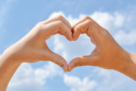 Hands forming a heart shape with blue sky and clouds in the backgroundの写真素材