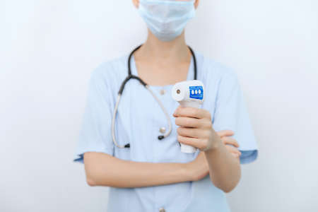 Temperature measurement gun in doctor hands. Close-up shot of doctor wearing protective surgical mask ready to use infrared isometric thermometer gun to check body temperature for virus symptomsの写真素材
