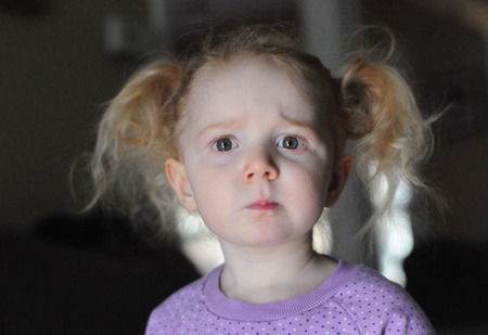 Portrait of an adorable blond little girl, with pigtails and a lavender T-shirt, looking at camera with a confused facial expression, indoorsの写真素材