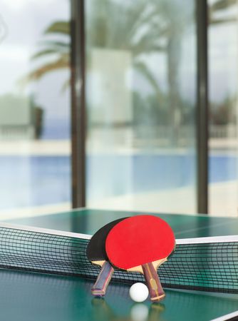Two table tennis or rackets and balls on a green table with net, swimming pool, palm tree and sea in the backgroundの写真素材