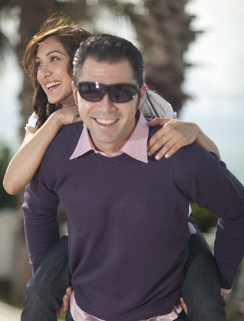 Young man piggybacking his girlfriend under palm tree smiling to the camera, selective focus on woman.の写真素材