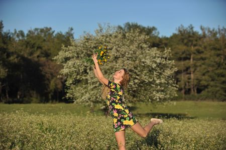Beautiful young blonde woman jumping in blooming meadow in spring, bunch of yellow flowers in hand; shallow depth of field, trees in backgroundの写真素材