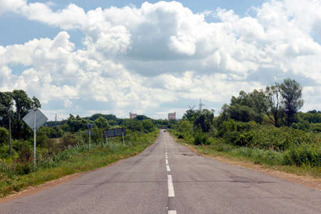 asphalt road highway going forward into perspective, passing by green fields against a blue sky with cloudsの写真素材