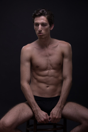 Nude young man sitting posing model. Caucasian skinny, slim, fit abs, tanned skin. Black background, low key.の写真素材