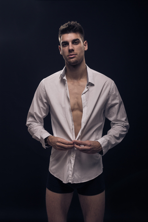 one young man buttoning white shirt, looking at camera, black background, underwearの写真素材