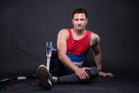 One disabled young adult man, prosthetic leg, athlete, sitting on floor, black backgroundの写真素材