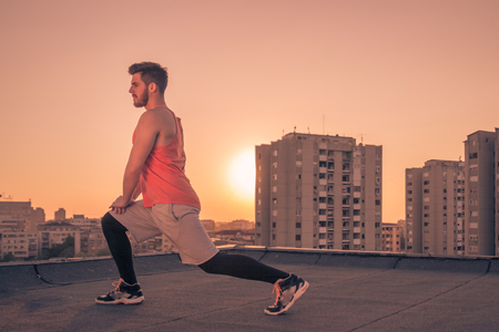 one young man only, athlete sport clothes, stretching leg, roof rooftop buildings behind, Sun sunset sunrise, warm colors, orange colorの写真素材
