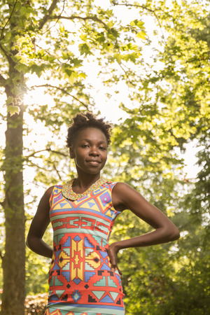 one, young adult, black african american woman 20-29 years, hands on hips, portrait sunny day outdoors, trees behind, low angle shot, wearing dressの写真素材