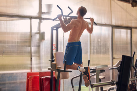one young man, stationary machine, pull-up exercise, legs extended. rear view.