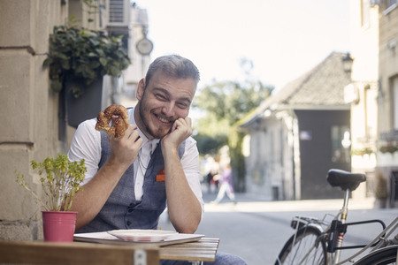 one young smiling man, 20-29 years old, wearing hipster suit, siting on street, eating pretzel in front of bakery outdoors. acting funny.の写真素材