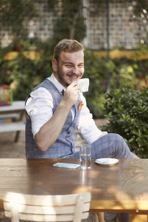 one young smiling man, 20-29 years old, wearing hipster suit, sitting at table in cafe garden, looking to camera, drinking espresso.の写真素材