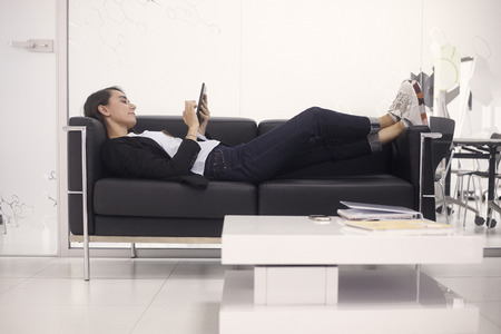 one young woman, white-collar worker, relaxing on sofa, working on tablet in modern office indoors.の写真素材