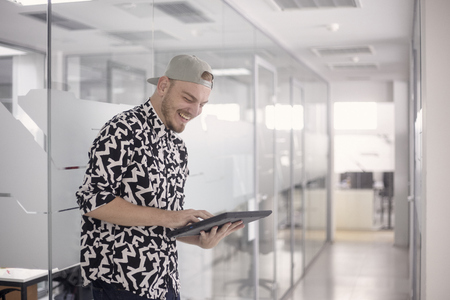 one young man, standing 20-29 years old, casual clothes, smiling laughing, using computer tablet. modern bright white office space.の写真素材