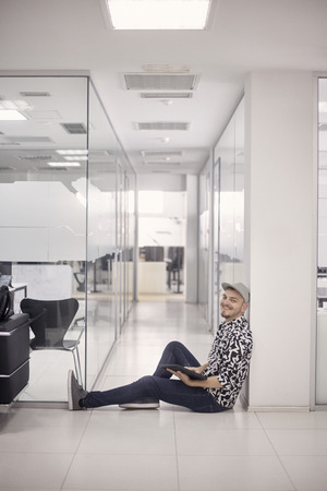 one young man smiling, 20-29 years old, sitting on floor, office space, modern interior. casual clothes, relaxing pose, looking at camera. holding laptop tablet.の写真素材
