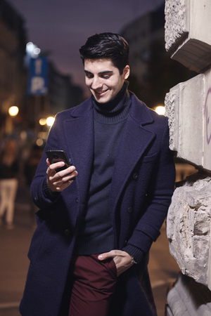 night, one handsome young man looking to smartphone, standing leaning on wall on street.の写真素材
