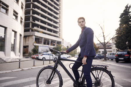 wide angle lens shot of city street buildings and cars, one man posing, sitting on bicycle outdoors. 20-29 years old, wearing suit. sunny day.の写真素材
