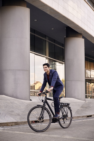 one young man, 20-29 years old, wearing suit, looking smiling. riding, pedaling standing, fancy  bicycle. full length body. modern architecture building behind.の写真素材