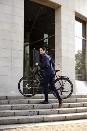 one young handsome man, wearing suit, pulling bicycle up on stairs steps, modern architecture building.の写真素材