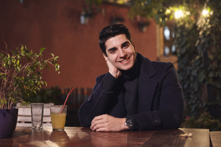 one young handsome man portrait, sitting in cafe garden at table, night sideways, smiling, looking to camera. upper body shot.の写真素材