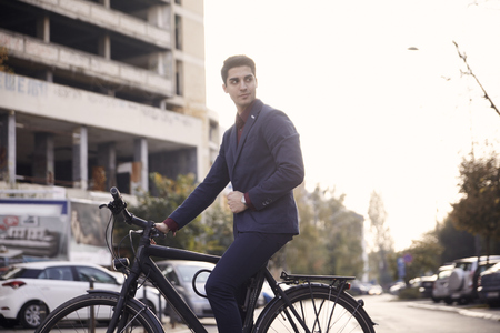wide angle lens shot of city street buildings and cars, one man posing, looking sideways,  sitting on bicycle outdoors. 20-29 years old, wearing suit. sunny day.の写真素材