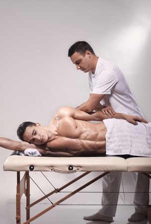 two young man, 20-29 years old, sports physiotherapy indoors in studio, photo shoot. Therapist masseur massaging arm of muscular patient laying on his side, with elbow.の写真素材