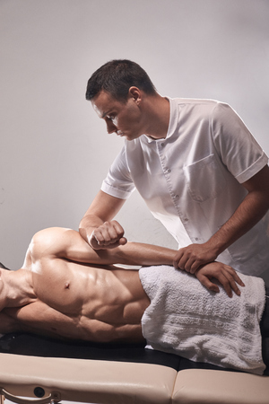 two young man, 20-29 years old, sports physiotherapy indoors in studio, photo shoot. Therapist masseur massaging arm of muscular patient laying on his side, with elbow.の写真素材