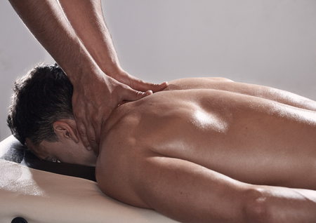 side view, two young man, 20-29 years old, sports physiotherapy indoors in studio, photo shoot. Physiotherapist massaging relaxed patient neck back side, with his hands.