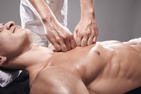side view, two young man, 20-29 years old, sports physiotherapy indoors in studio, photo shoot. Masseur massaging athlete mans chest, muscular body, hands close-up.の写真素材