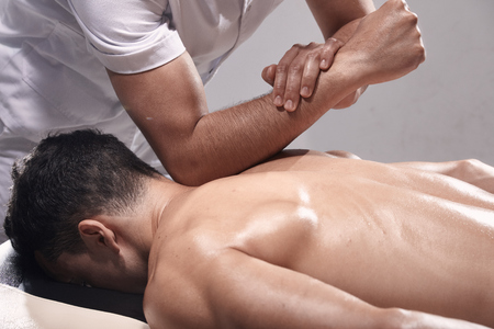 side view, two young man, 20-29 years old, sports physiotherapy indoors in studio, photo shoot. Strong Physiotherapist hard massaging relaxed patient neck back side, with his elbow.の写真素材